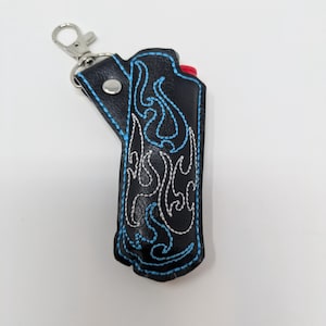 May include: Black leather lighter case with blue and white flame embroidery. The case has a silver-colored metal clasp and a snap closure. The lighter's red button is visible. A stylish accessory.