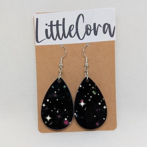 May include: Teardrop-shaped earrings with a black background and a galaxy design featuring white stars, pink, and green specks. The earrings hang from silver-colored hooks and are displayed on a brown card with the text "LittleCora" in a handwritten font.