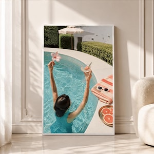 Op de afbeelding: Een ingelijste kunstdruk met een persoon in een turquoise badpak in een zwembad, die een roze hibiscusbloem en een cocktail vasthoudt. Naast het zwembad liggen een gestreepte handdoek, een witte zonnebril en een bord met schijfjes grapefruit.