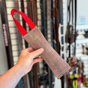 May include: A rectangular dog tug toy made of a textured, weathered brown fabric with a prominent red nylon strap handle attached at the top. The toy features visible white stitching along the edges and a worn, mottled surface texture throughout.