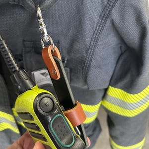Pode incluir: Um rádio bidirecional amarelo e preto preso a uma jaqueta cinza escuro com listras refletivas amarelas. O rádio é preso com um clipe de couro e metal.