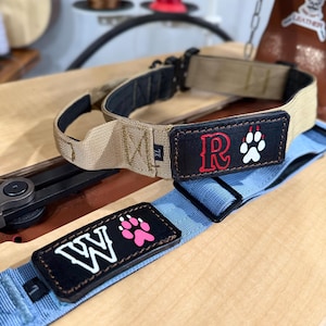 May include: A tan dog collar with a black leather patch featuring a red letter R and a white paw print, and a light blue dog collar with a black leather patch featuring a white letter W and a pink paw print, both with black plastic buckles.