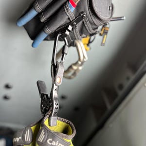 May include: A black metal spring clamp with a leather strap and a carabiner attached to a tool pouch. The clamp holds a pair of lime green and grey work gloves. The clamp features a cutout heart pattern on its handle and a copper-coloured rivet.