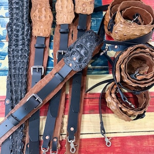 Könnte beinhalten: Eine Sammlung von Gitarrengurten mit strukturierten, geprägten Lederpolstern in Schwarz und Hellbraun, kombiniert mit glatten schwarzen Lederverstellteilen, silberfarbenen Metallschnallen und drehbaren Karabinerhaken zur Befestigung an Instrumenten.
