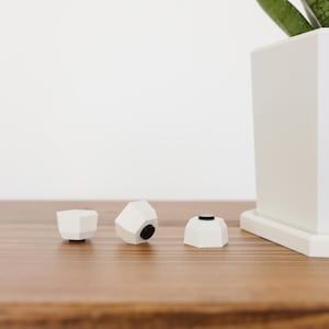 May include: Three white, geometric bottle stoppers with black accents are displayed on a wooden surface. The stoppers have a modern, minimalist design. A white square planter with a green plant is in the background.