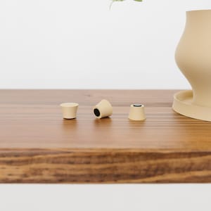 May include: Three beige bottle stoppers with black interiors are arranged on a wooden surface. A beige ceramic planter with a matching saucer is in the background. The stoppers are cylindrical and conical.