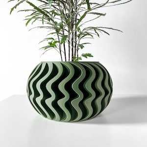 May include: A round, olive green planter featuring a repeating vertical wave pattern along its exterior surface. The planter has a wide, open top and contains a leafy green plant with thin, dark stems extending upward from the center.