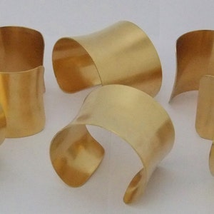 May include: Six gold-colored metal cuff bracelets. The bracelets have a curved, open design. The metal has a smooth, polished finish, reflecting light. The cuffs are arranged on a white surface, showcasing their shape and color.