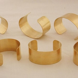 May include: Six gold-colored cuff bracelets. The bracelets have a simple, open design with a curved shape. The metal has a smooth, polished finish. The bracelets are displayed on a light-colored surface.