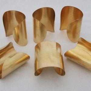 May include: Six gold metal napkin rings, each with a unique curved design. The rings are arranged in a semi-circle on a white surface.