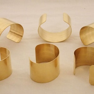 May include: Six gold-toned metal bracelets, each with a slightly different shape and width. The bracelets are arranged in a semi-circle on a white surface.