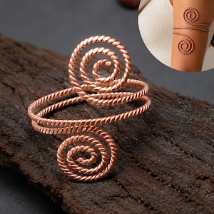 May include: Copper-coloured arm cuff with a spiral design. The cuff features two spiral accents and a twisted wire design. The jewellery is displayed on a dark wooden surface. The cuff is a unique accessory.