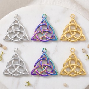 Puede incluir: Seis colgantes de nudo celta en tonos plateado, iridiscente y dorado. Cada colgante presenta un diseño triangular con líneas entrelazadas, adecuado para la fabricación de joyas o manualidades. Los colgantes se exhiben sobre una superficie de mármol blanco.