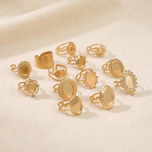 Puede incluir: Una colección de anillos dorados con varios diseños. Algunos anillos presentan engastes ovalados o redondos, mientras que otros tienen patrones de rayos de sol o texturizados. Los anillos se exhiben sobre una superficie de color crema, mostrando sus intrincados detalles.