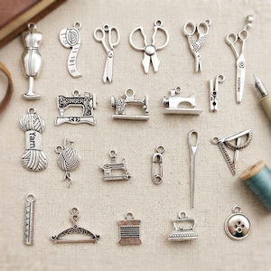 May include: A collection of silver-toned sewing-themed charms. The charms include scissors, sewing machines, measuring tapes, dress forms, yarn, needles, and buttons. The word "SINGER" is visible on one charm, and "Yarn" on another. The charms are arranged on a neutral background.