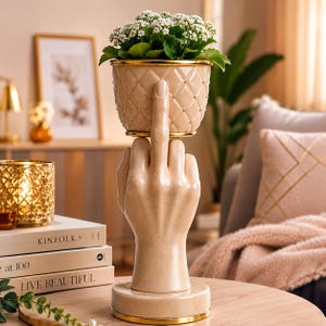 May include: A beige ceramic planter featuring a quilted diamond pattern with gold accents, held up by a sculpted hand base with an extended middle finger. The planter contains a small green plant with white flowers, creating a unique decorative piece.