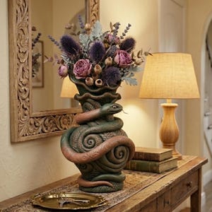May include: A decorative vase featuring a green and brown snake design, holding a bouquet of purple and pink flowers. The vase is on a wooden table with a gold tray and a lamp.