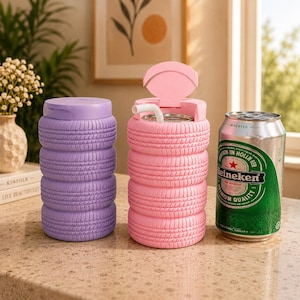 May include: Two insulated tumblers, one lavender and one pink, shaped like stacked tires. The pink tumbler has a lid and straw. A Heineken beer can is next to the pink tumbler. The tumblers are on a light-colored surface.