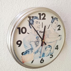 Peut inclure: Une horloge murale ronde avec un cadre argenté et un cadran crème. Le cadran présente de grands chiffres noirs et une illustration de trois personnes. Le texte "Flash" et "CHIPIE" est imprimé sur le cadran, avec "MADE IN FRANCE".