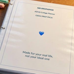 Könnte beinhalten: Ein offener weißer Ordner auf einer Holzoberfläche mit dem Text "NEUROPAPER", "ADHD College Planner" und "EXAM PREP PACK". Ein blaues Herzsymbol ist vorhanden. Der Satz "Made for your real life, not your ideal one" ist darunter gedruckt.