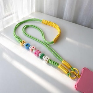 May include: A phone lanyard featuring a green cord with white stripes, accented by yellow braided sections and a series of colorful letter beads spelling out the name SOPHIE, interspersed with small white round beads, attached to a pink phone case.