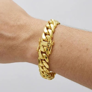 May include: A gold-colored curb chain bracelet with a secure clasp. The bracelet is made of interlocking links and is worn on a wrist. The links are thick and have a polished finish.