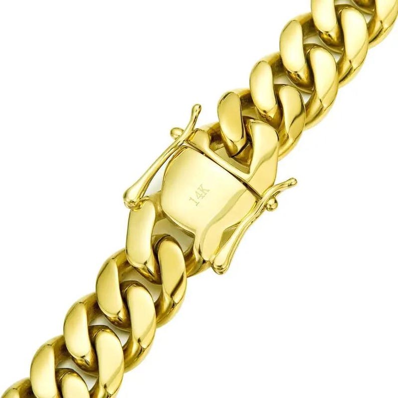 May include: Close-up of a gold-colored curb chain necklace with a secure clasp. The clasp is engraved with "14K". The chain links are thick and rounded, reflecting light. The jewelry is set against a white background.