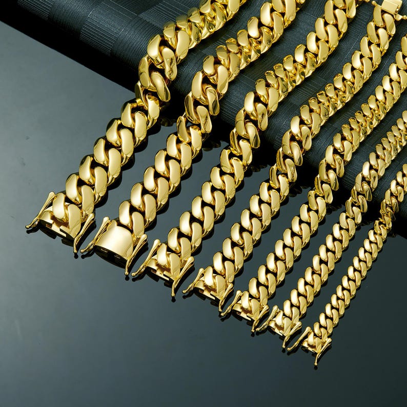 May include: A collection of gold-colored, curb-link chain necklaces and bracelets. The jewelry pieces are arranged diagonally, showcasing varying widths and clasp styles. The chains have a polished, reflective surface, and are displayed against a dark background.