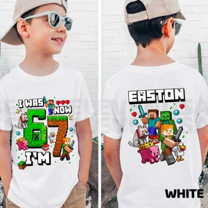 May include: A white t-shirt featuring Minecraft-themed graphics. The front displays the text I WAS NOW 67 I'M with a large green number 6 and brown number 7, surrounded by game characters. The back shows the name EASTON above game characters.