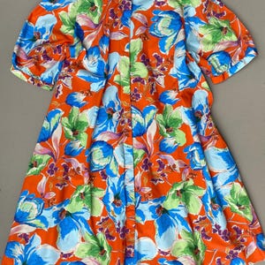Könnte beinhalten: Ein kurzärmeliges Hemd mit durchgehender Knopfleiste auf einem leuchtend orangefarbenen Hintergrund mit einem großflächigen Blumenmuster in leuchtendem Blau, Hellgrün und Violett. Das Kleidungsstück hat einen klassischen Kragen und eine lockere Passform.