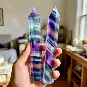 May include: Two polished fluorite crystal towers featuring distinct banded patterns of deep purple, teal, and clear white. Each hexagonal point displays natural colour zoning and translucent layers, showcasing a smooth, faceted finish throughout.
