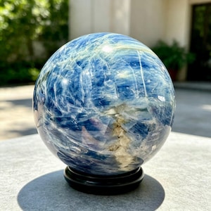 Puede incluir: Una esfera de cianita azul pulida con patrones veteados en azul oscuro y claro, junto con inclusiones cristalinas blancas. La piedra lisa y esférica descansa sobre un soporte de exhibición negro, circular y de diseño sencillo.