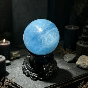 Puede incluir: Una bola de cristal esférica de color azul claro pulido con vetas e inclusiones internas visibles, que descansa sobre una base de pedestal circular oscura y ornamentada. La superficie de la esfera refleja la luz, resaltando su forma.