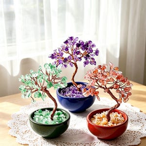 May include: Three decorative wire bonsai trees featuring polished gemstone chips as leaves. One tree has green stones in a green bowl, one has purple stones in a blue bowl, and one has orange stones in a red bowl, all with twisted copper-colored wire trunks.