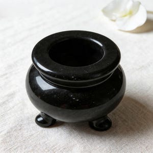 May include: A small, polished black stone cauldron with a smooth, rounded body and a wide, circular opening at the top. The vessel is supported by three short, sturdy, rounded feet, providing a stable base for this solid, dark-colored container.