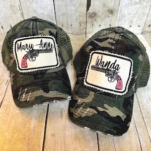 May include: Two camouflage trucker hats with a distressed look. Each hat has a patch with a gun and a name. One hat says "Mary-Anne" and the other says "Wanda".