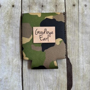 May include: A green, brown, and black camouflage can cooler with a brown paper tag that says "Goodbye Earl".