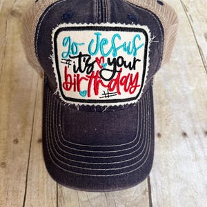 May include: A black and brown trucker hat with a distressed denim patch that says "Go Jesus it's your birthday" in colourful embroidery.