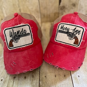 May include: Two red trucker hats with distressed detailing. Each hat features a patch with a revolver graphic and a name: "Wanda" and "Mary Ann". The hats have a tan mesh back and a curved brim.