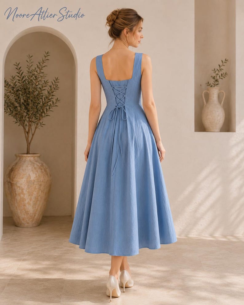 May include: A cornflower blue midi dress featuring a square neckline, wide shoulder straps, and a lace-up corset-style back closure with a long tie. The dress has a fitted bodice and a full, flared skirt that falls below the knee.