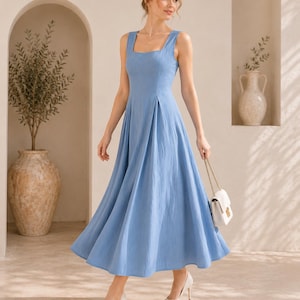 May include: A light blue midi dress with a square neckline, wide shoulder straps, and a pleated skirt. The dress is paired with white pointed-toe heels and a small white handbag with a gold chain strap. The text Noore Attier Studio is visible.