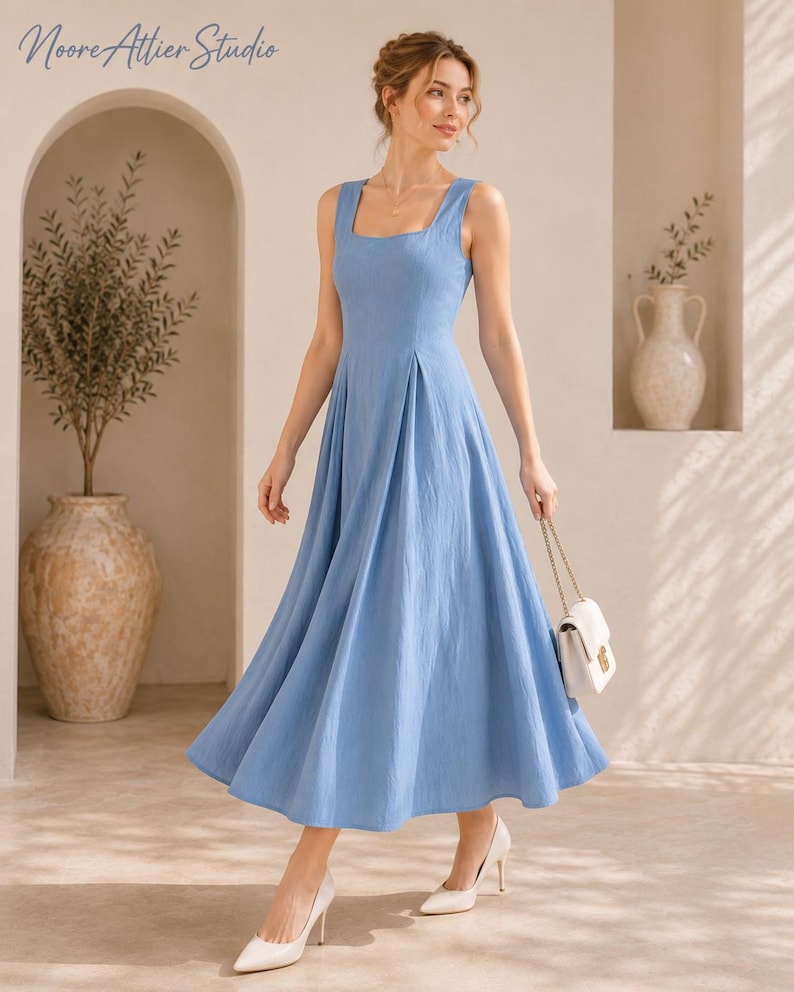 May include: A light blue midi dress with a square neckline, wide shoulder straps, and a pleated skirt. The dress is paired with white pointed-toe heels and a small white handbag with a gold chain strap. The text Noore Attier Studio is visible.