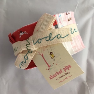 May include: A set of fabric scraps for quilting, featuring a pink and white striped pattern, a red fabric with white dog silhouettes, and a white fabric with a girl on a swing. The fabric scraps are tied together with a white ribbon with the word "moda" written in green. The label reads "sherbet pips by Aneela Hoey moda".