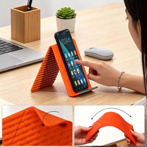 May include: An orange and gray phone stand holds a smartphone displaying an app screen. The stand is made of flexible material and has a textured surface. The stand is on a wooden desk with a laptop, mouse, and small plant.