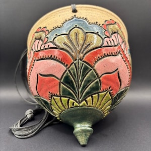 May include: A hanging ceramic planter with a rounded, tapered base and a speckled beige rim. The body features deeply carved, raised floral patterns glazed in vibrant shades of coral, pink, forest green, and muted blue, suspended by a grey cord.