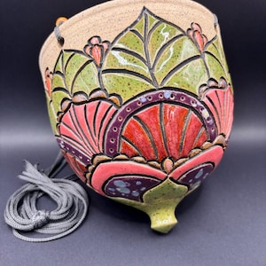 May include: A ceramic hanging planter with a speckled beige base featuring a hand-carved, relief-style pattern of green leaves and coral-pink fan shapes outlined in dark purple. The bowl-shaped vessel includes a pointed bottom and grey cord.