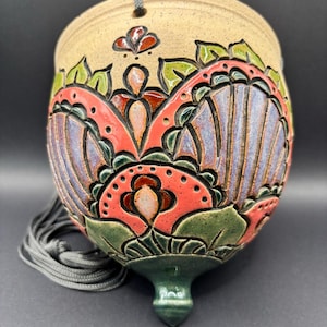 May include: A hanging ceramic planter with a speckled beige base and a carved, relief-style floral pattern. The design features vibrant red, purple, and green glazes. It includes a pointed bottom finial and a grey cord for suspension.