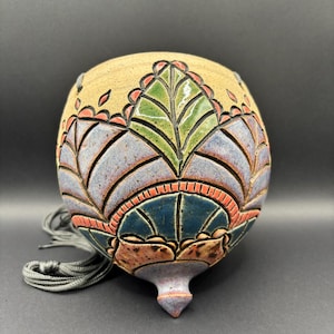 May include: A round, hanging ceramic planter with a beige base and a carved, multi-colored leaf pattern in shades of green, blue, and orange. The vessel features a pointed bottom and includes grey hanging cords attached to the top rim.