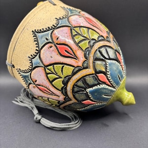 May include: A ceramic hanging planter with a speckled beige base and a carved, multi-colored floral pattern in shades of pink, green, and blue. The vessel features a pointed bottom and includes a grey cord for suspension, showcasing a textured finish.