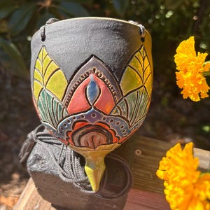 May include: A hanging ceramic planter with a matte black exterior featuring a hand-painted, colorful lotus-inspired design in shades of yellow, green, orange, blue, and purple. The vessel has a pointed base and includes grey hanging cords.